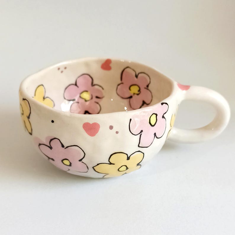 Może przedstawiać: Ręcznie robiony ceramiczny kubek z białą podstawą, ozdobiony r&oacute;żowymi i ż&oacute;łtymi wzorami kwiat&oacute;w obrysowanymi na czarno. Kubek ma zaokrąglony kształt, uchwyt i małe r&oacute;żowe akcenty w kształcie serca. Idealny do kawy lub herbaty.
