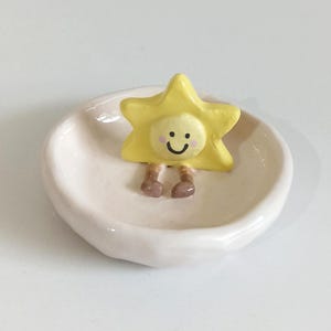 Puede incluir: Un plato de cerámica blanco con una figurita amarilla en forma de estrella con una cara sonriente. La figurita tiene piernas marrones y está sentada en el plato.