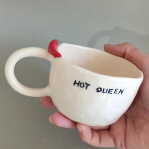 May include: White ceramic mug with a large handle and the words "HOT QUEEN" in black script. A red chili pepper with a green stem adorns the rim. The mug is designed for beverages.