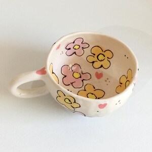 Może przedstawiać: Mały, kremowy ceramiczny kubek z uchwytem, ​​ozdobiony r&oacute;żowymi i ż&oacute;łtymi kwiatami obrysowanymi na czarno. Wnętrze ma ten sam kwiatowy wz&oacute;r, wraz z małymi r&oacute;żowymi sercami. Kubek jest przeznaczony do napoj&oacute;w.