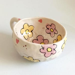 Może przedstawiać: Mała, kremowa ceramiczna miska z uchwytem, ​​ozdobiona ręcznie malowanymi ż&oacute;łtymi i r&oacute;żowymi kwiatami, małymi r&oacute;żowymi sercami i brązowymi kropkami. Miska ma zaokrąglony kształt i gładkie, błyszczące wykończenie.