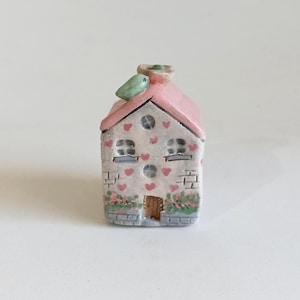 Puede incluir: Una casa de cerámica en miniatura con techo rosa, paredes blancas y una pequeña chimenea. La casa está decorada con corazones rojos, pequeñas ventanas y una puerta de madera. El follaje verde acentúa la base de la casa.