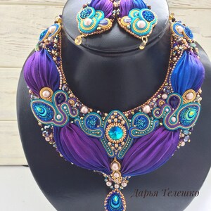 May include: A purple and blue beaded necklace with a teardrop pendant and matching earrings. The necklace features a gold and turquoise design with pearls and crystals. The earrings are also decorated with pearls and crystals. The necklace and earrings are on a black display stand. "Дарья Телешко" is written in the bottom right corner of the image.