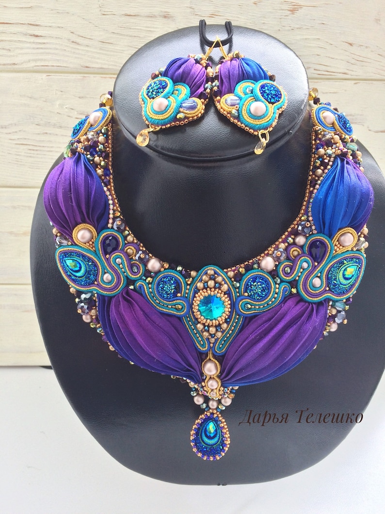 May include: A purple and blue beaded necklace with a teardrop pendant and matching earrings. The necklace features a gold and turquoise design with pearls and crystals. The earrings are also decorated with pearls and crystals. The necklace and earrings are on a black display stand. "Дарья Телешко" is written in the bottom right corner of the image.