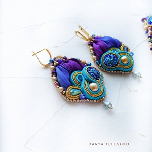 May include: A pair of dangle earrings with a teardrop shape. The earrings are made with blue, purple, and gold fabric, beads, and glitter. The earrings have a gold hook closure.
