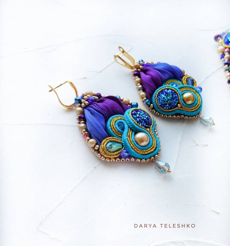 May include: A pair of dangle earrings with a teardrop shape. The earrings are made with blue, purple, and gold fabric, beads, and glitter. The earrings have a gold hook closure.