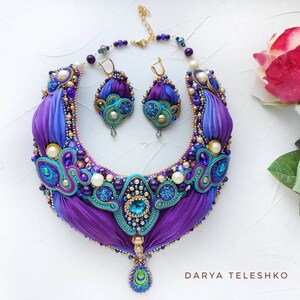 May include: A purple and blue silk ribbon necklace with gold accents and pearl beads. The necklace has a matching pair of earrings with a similar design. The necklace and earrings are on a white background.