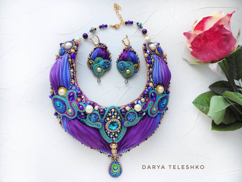 May include: A purple and blue silk ribbon necklace with gold accents and pearl beads. The necklace has a matching pair of earrings with a similar design. The necklace and earrings are on a white background.