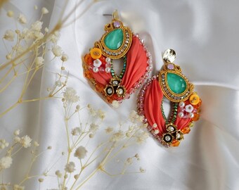May include: A pair of handcrafted statement earrings. Each earring features a large, teardrop-shaped, light green gemstone, surrounded by gold-toned metalwork and embellished with orange fabric, beads, and pearls. The earrings are set against a white satin background.