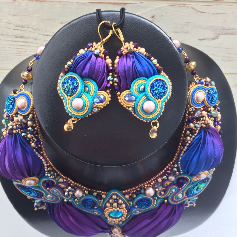 May include: A purple and blue silk ribbon necklace and matching earrings with gold accents and pearl-like beads. The jewelry is displayed on a black stand.