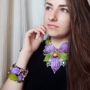 May include: A woman wearing a black top with a large purple and green flower-shaped necklace and a matching bracelet. The necklace and bracelet are made of fabric and beads.