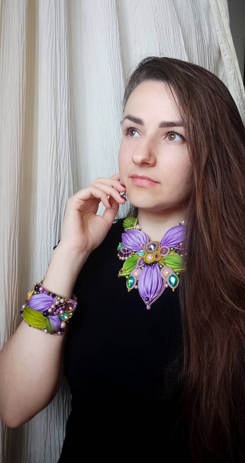 May include: A woman wearing a black top with a large purple and green flower-shaped necklace and a matching bracelet. The necklace and bracelet are made of fabric and beads.