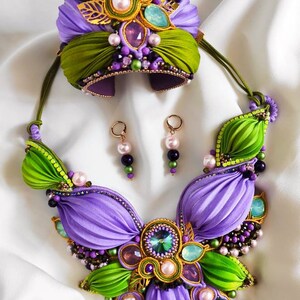 May include: A purple and green jewelry set with a necklace, bracelet, and earrings. The necklace and bracelet feature a flower design with green and purple fabric, pearls, and crystals. The earrings are gold with purple and green beads.