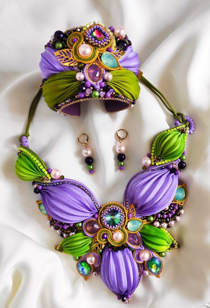 May include: A purple and green jewelry set with a necklace, bracelet, and earrings. The necklace and bracelet feature a flower design with green and purple fabric, pearls, and crystals. The earrings are gold with purple and green beads.