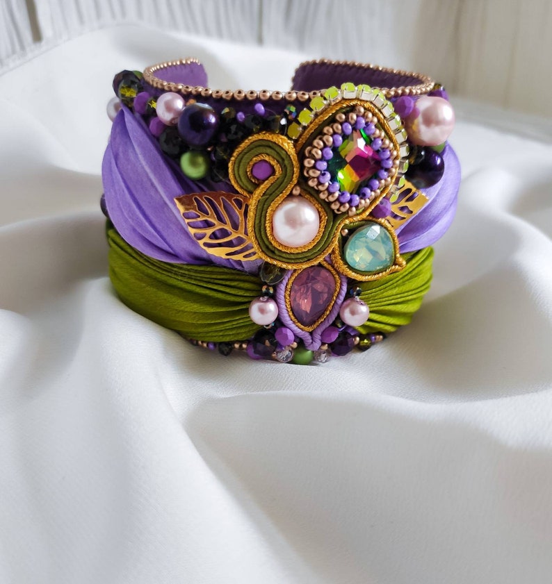May include: A purple and green fabric bracelet with a gold leaf design and various colored beads and gems.