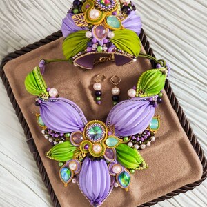 May include: A set of purple and green jewelry featuring a necklace, bracelet, and earrings. The necklace and bracelet are made of fabric and decorated with pearls, beads, and crystals. The earrings are made of pearls and beads.