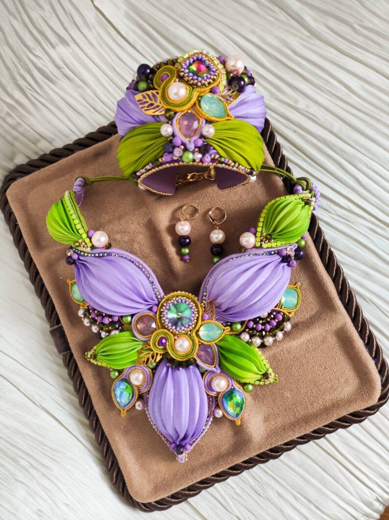 May include: A set of purple and green jewelry featuring a necklace, bracelet, and earrings. The necklace and bracelet are made of fabric and decorated with pearls, beads, and crystals. The earrings are made of pearls and beads.