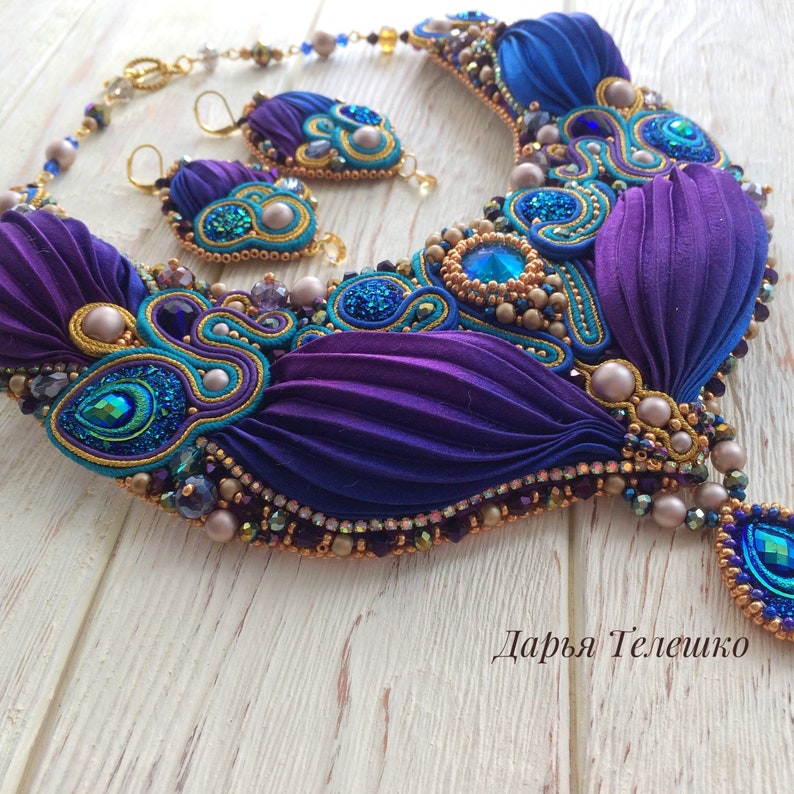 May include: A purple, blue, and teal silk ribbon necklace with gold accents and crystal beads. The necklace is paired with matching earrings. The necklace is on a white wooden surface. "Дарья Телешко" is written in black text on the surface.