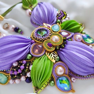 May include: A purple and green flower-shaped necklace with gold accents and various beads. The necklace is made of fabric and features a variety of crystals and pearls.