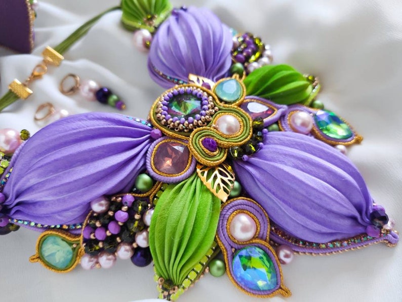 May include: A purple and green flower-shaped necklace with gold accents and various beads. The necklace is made of fabric and features a variety of crystals and pearls.