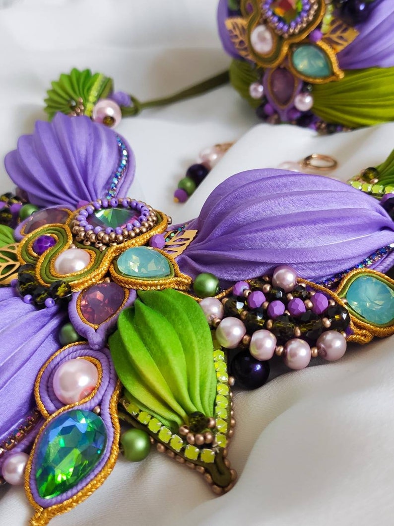 May include: A purple and green beaded necklace with iridescent gemstones and pearls. The necklace is made with fabric flowers and gold trim.