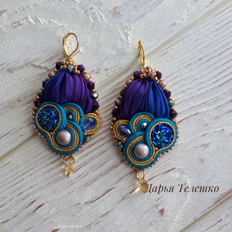 May include: Purple and blue silk ribbon earrings with gold accents and sparkly blue beads. The earrings are teardrop shaped and have a gold hook.