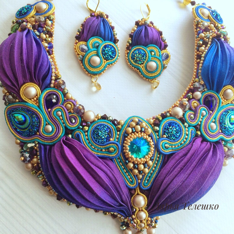 May include: A purple and blue statement necklace with gold and teal accents. The necklace is made of fabric and features a variety of beads and crystals. Matching earrings are also included.