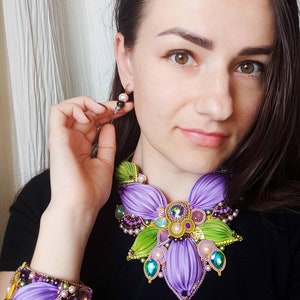 May include: A purple and green flower-shaped necklace and matching bracelet with beaded accents. The jewelry is made of fabric and features a variety of colors, including purple, green, gold, and pink.