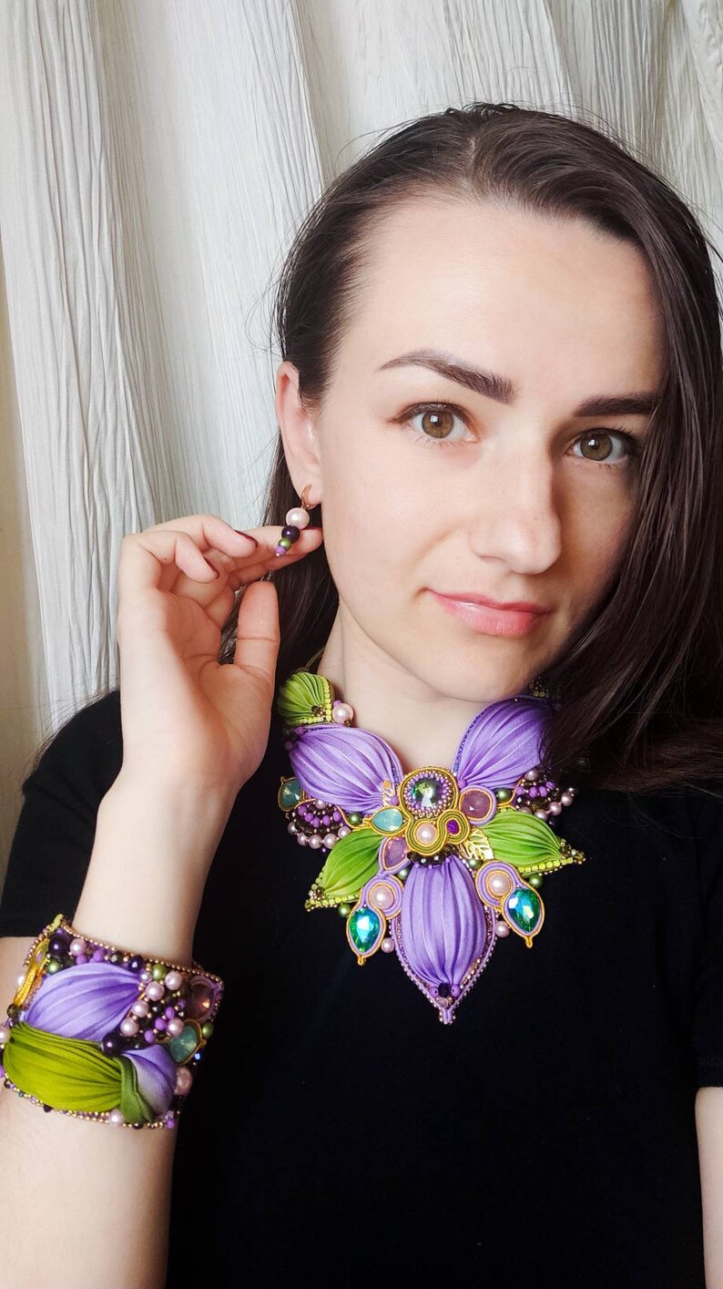 May include: A purple and green flower-shaped necklace and matching bracelet with beaded accents. The jewelry is made of fabric and features a variety of colors, including purple, green, gold, and pink.