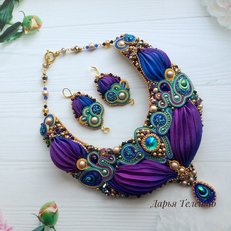May include: A purple and blue silk ribbon necklace with gold accents and a matching pair of earrings. The necklace features a large, ornate pendant with a teardrop-shaped design. The earrings are smaller and have a similar design. The jewelry is decorated with pearls, crystals, and glitter.