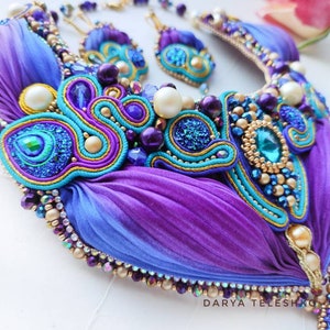May include: A purple and blue silk ribbon necklace with gold and turquoise embellishments. The necklace features a large, ornate centerpiece with a turquoise gemstone and smaller, iridescent beads. The necklace is accented with gold and silver beads and a delicate chain.