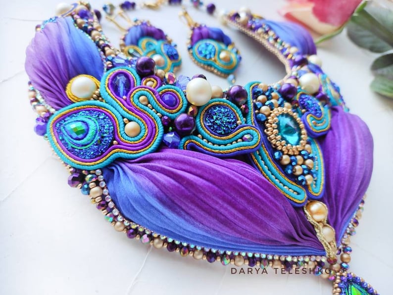May include: A purple and blue silk ribbon necklace with gold and turquoise embellishments. The necklace features a large, ornate centerpiece with a turquoise gemstone and smaller, iridescent beads. The necklace is accented with gold and silver beads and a delicate chain.