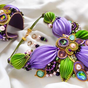 May include: A purple and green silk flower necklace with gold accents and crystal embellishments. The necklace is paired with matching earrings and a bracelet.