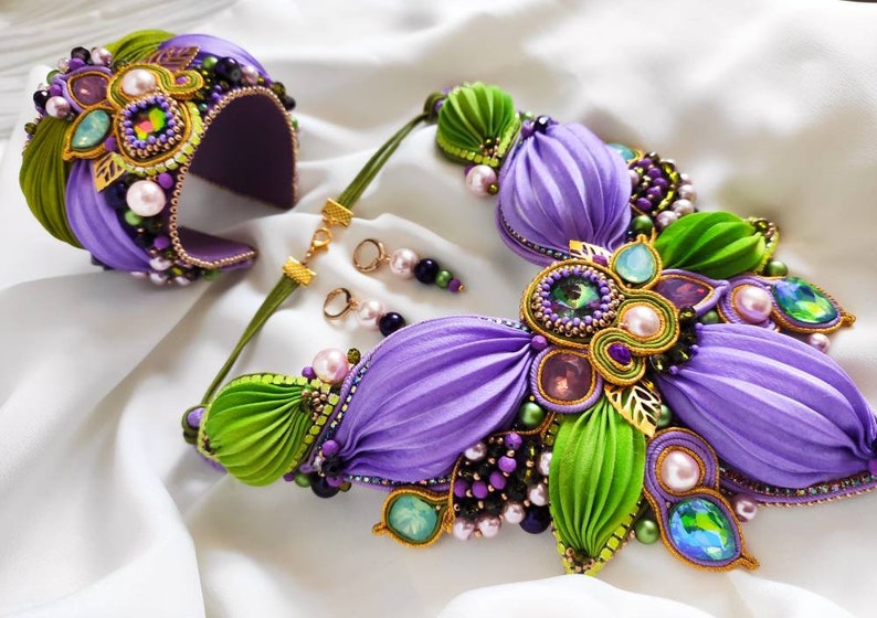 May include: A purple and green silk flower necklace with gold accents and crystal embellishments. The necklace is paired with matching earrings and a bracelet.
