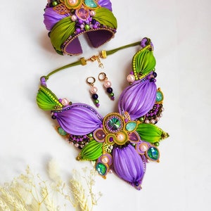 May include: A purple and green silk ribbon jewelry set with a statement necklace, bracelet, and earrings. The necklace features a large flower design with green leaves and purple petals. The bracelet is a wide cuff with a similar design. The earrings are small and dangle.