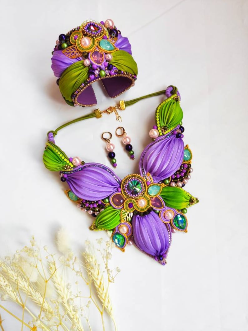 May include: A purple and green silk ribbon jewelry set with a statement necklace, bracelet, and earrings. The necklace features a large flower design with green leaves and purple petals. The bracelet is a wide cuff with a similar design. The earrings are small and dangle.