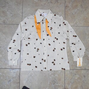 Op de afbeelding: Een wit overhemd met lange mouwen met een zwarte en oranje print. Het shirt heeft een kraag en een knoopsluiting aan de voorkant. Het shirt heeft een gele strik.