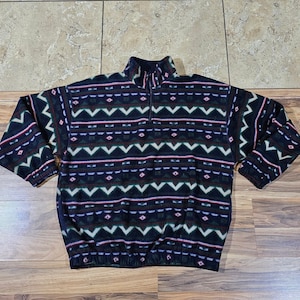 May include: A dark blue fleece pullover with a geometric pattern in white, green, and red. The long-sleeved top has a quarter-zip closure at the neck and a gathered hemline. The pattern includes zigzags, diamonds, and horizontal lines.