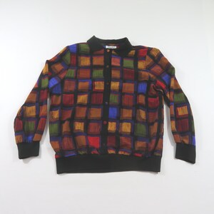 May include: A long-sleeve button-up shirt featuring a multi-colored grid pattern of squares in shades of brown, blue, red, and green. The garment includes a solid black collar, black cuffs, a black hem, and a black vertical button placket.