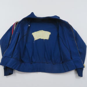 May include: A blue zip-up track jacket featuring a high collar and long sleeves with red and white stripes along the shoulders. The interior lining is blue and displays a central, rectangular, cream-colored fabric patch with frayed edges.