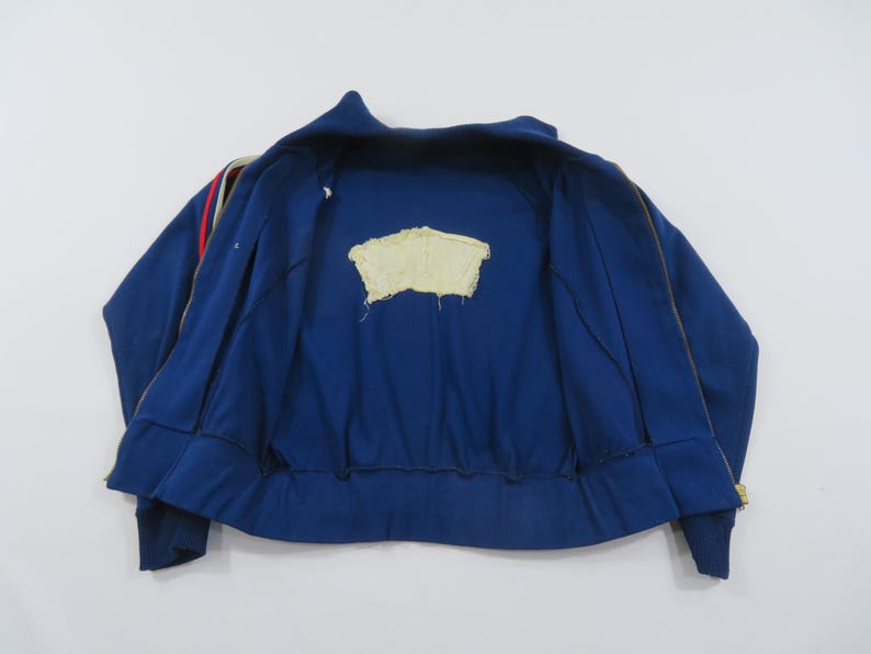 May include: A blue zip-up track jacket featuring a high collar and long sleeves with red and white stripes along the shoulders. The interior lining is blue and displays a central, rectangular, cream-colored fabric patch with frayed edges.