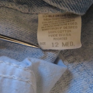 May include: A white care label with blue text that reads "Tumble Dry Med", "Wash Dark and Light Colors Separate", "Only Non-Chlorine Bleach When Needed", "Warm Iron If Desired", "100% Cotton", "Made in USA", "RN34783", "Size 12 Med."