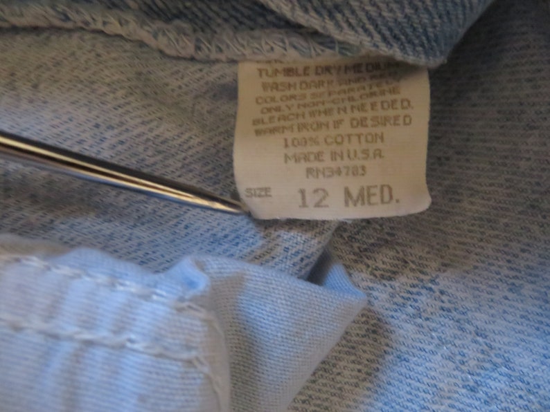 May include: A white care label with blue text that reads "Tumble Dry Med", "Wash Dark and Light Colors Separate", "Only Non-Chlorine Bleach When Needed", "Warm Iron If Desired", "100% Cotton", "Made in USA", "RN34783", "Size 12 Med."