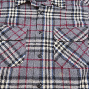 May include: A long-sleeve button-down shirt featuring a gray, black, white, and red plaid pattern. The shirt includes a pointed collar and two chest pockets with flaps, each secured with a button. The front placket is fastened with dark buttons.