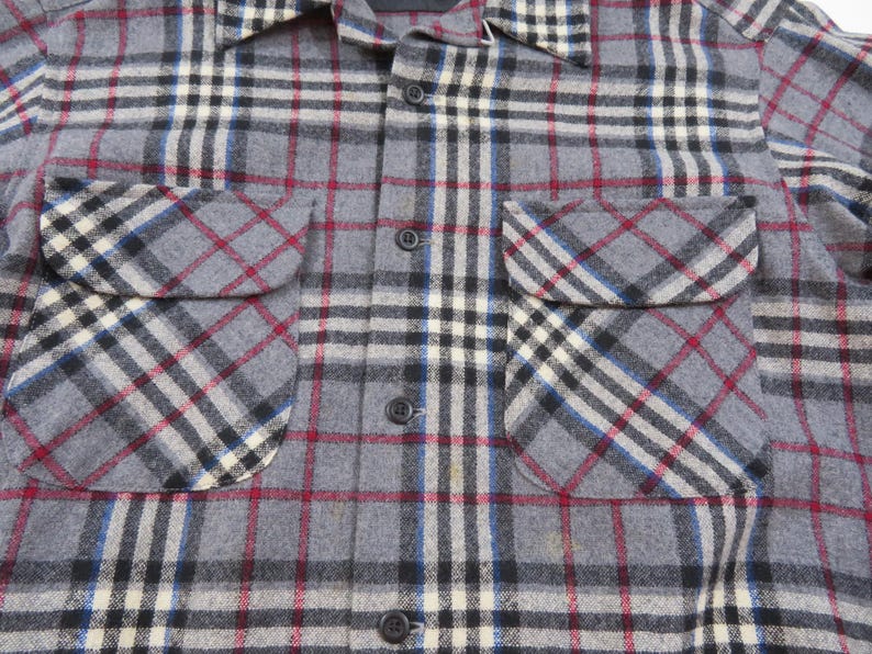 May include: A long-sleeve button-down shirt featuring a gray, black, white, and red plaid pattern. The shirt includes a pointed collar and two chest pockets with flaps, each secured with a button. The front placket is fastened with dark buttons.
