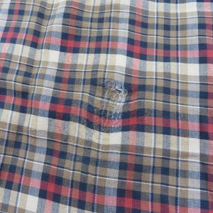 May include: A woven fabric featuring a plaid pattern in shades of tan, navy blue, and red. The material displays a visible area of wear and fraying near the center, where the threads have become loose and thin, disrupting the consistent grid design.