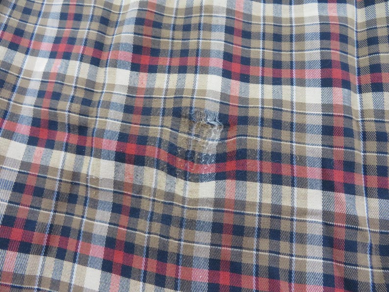 May include: A woven fabric featuring a plaid pattern in shades of tan, navy blue, and red. The material displays a visible area of wear and fraying near the center, where the threads have become loose and thin, disrupting the consistent grid design.