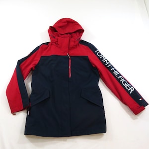 May include: A navy blue and red hooded jacket featuring a front zipper closure, two flap pockets, and the text TOMMY HILFIGER printed in white along the left sleeve. The jacket has a color-blocked design with red on the hood and upper sleeves.