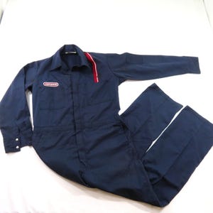 May include: A dark blue long-sleeve jumpsuit featuring a collar, buttoned cuffs, and two chest pockets. The garment displays a red and white vertical stripe on the left shoulder and a Conoco logo patch on the right side of the chest area.