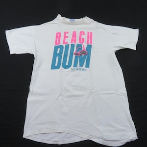 Puede incluir: Camiseta blanca con las palabras "BEACH BUM NEWPORT" en rosa y azul. El diseño incluye gafas de sol. La camiseta está colocada sobre una superficie negra.